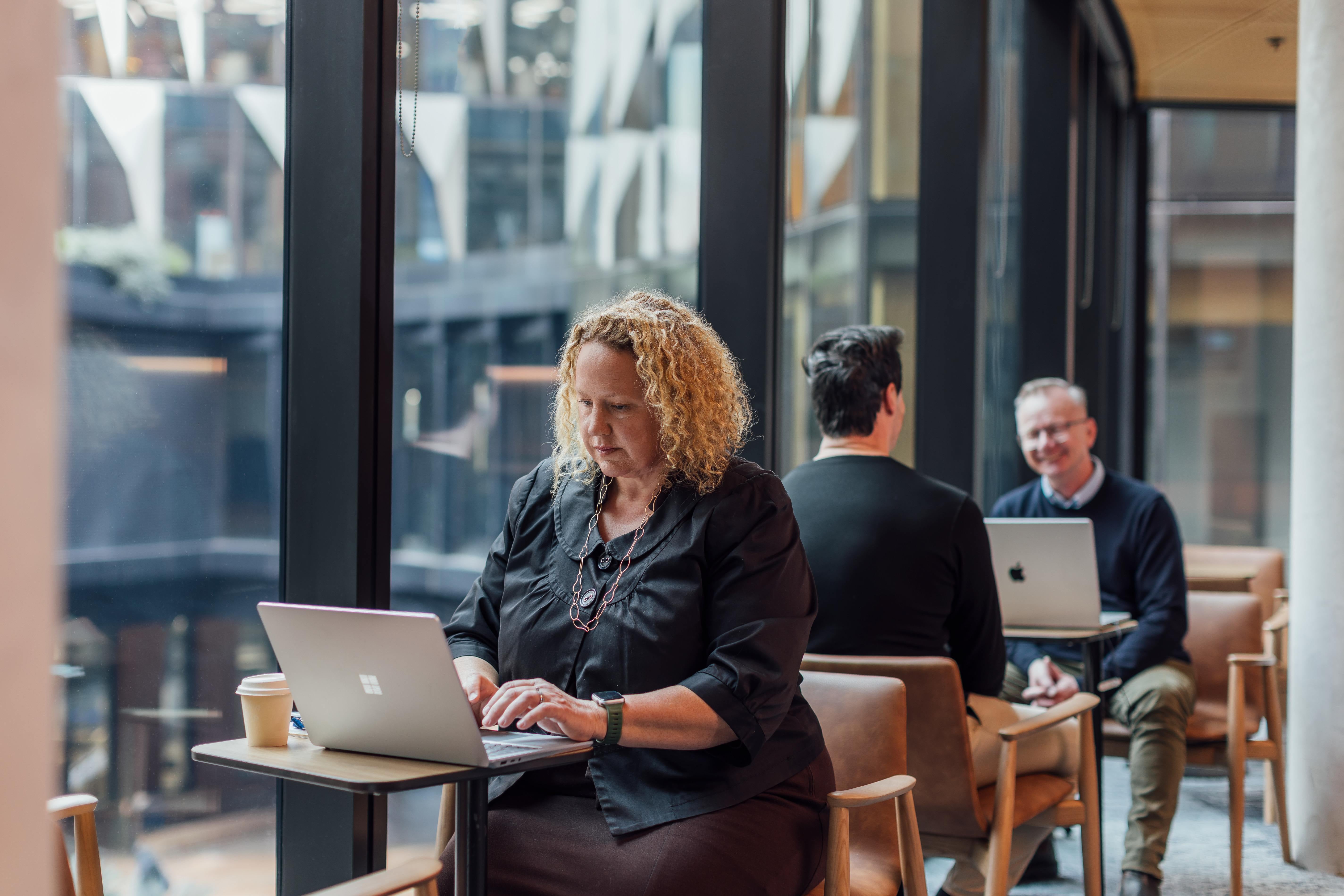 A woman with curly hair works on a laptop at a café table, with two other people seated nearby and large windows in the background.