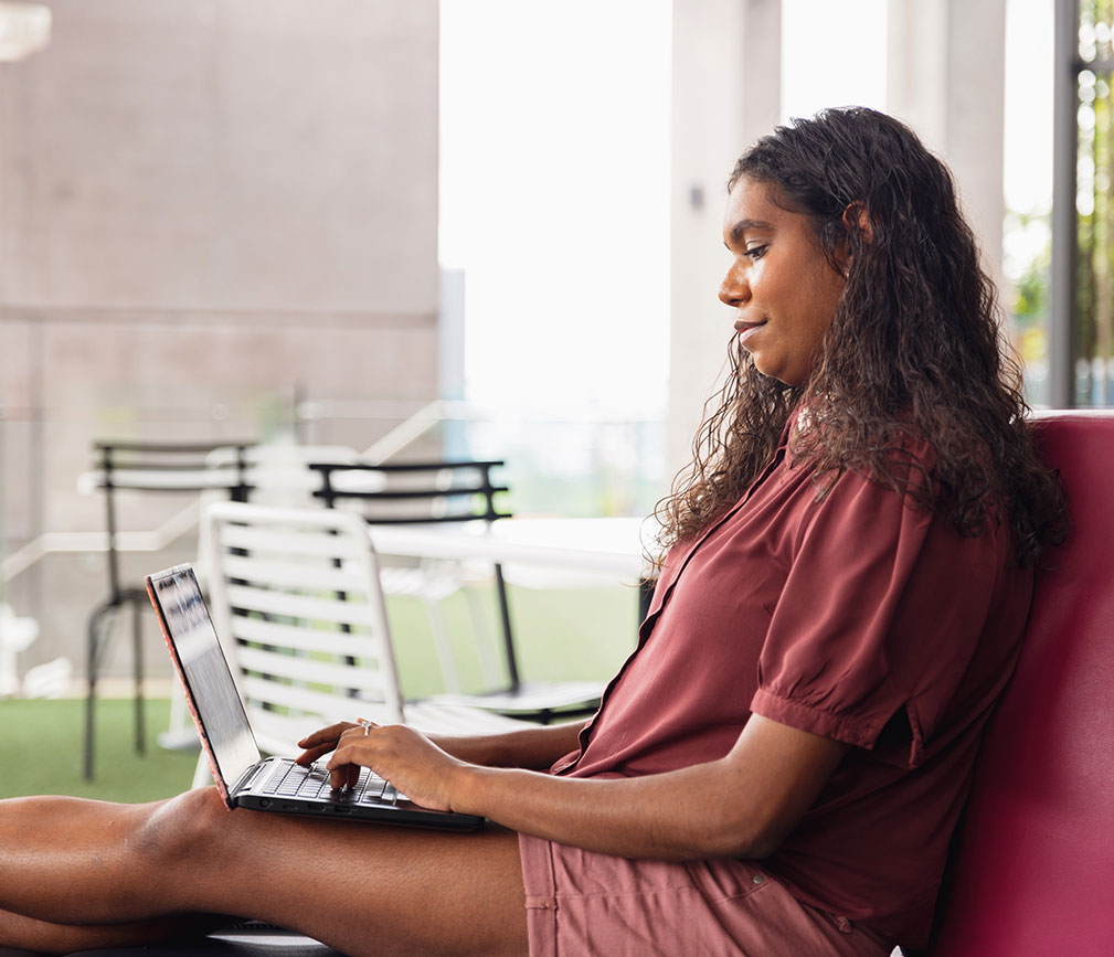 A woman with dark hair reclines on a plum couch with a laptop in her lap. She appears focused on what she's typing.