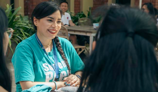 A woman in a teal t-shirt smiling at someone in an outdoor setting.
