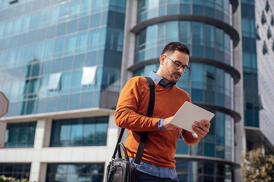 A man with dark hair and glasses smiles at a digital tablet he is holding. He is outdoors and may be on his way to work or university.