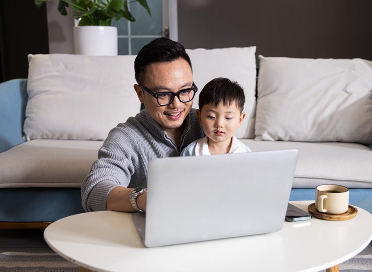 Man with glasses and his young son sitting on the ground, looking and pointing at his laptop.