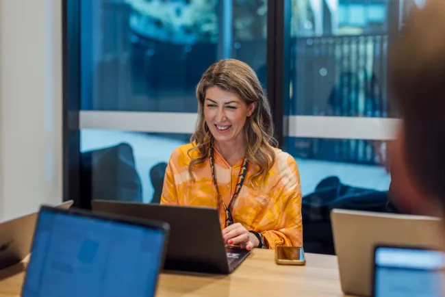 A woman in an orange tie-dye shirt smiles while working at a desk with a laptop, tablet, and smartphone in a modern office with large windows and blurred colleagues in the background.