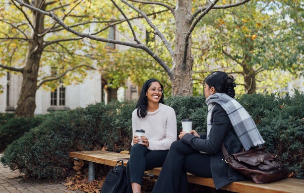 Two women engaged in conversation while sitting on a bench in a park setting and drinking coffee.