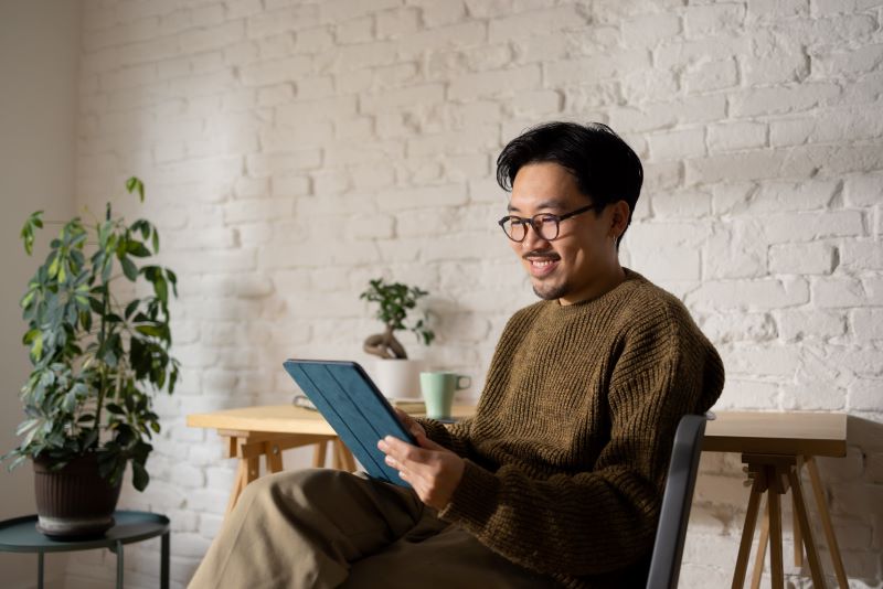 A man wearing glasses sits on a chair, interacting with a tablet device.